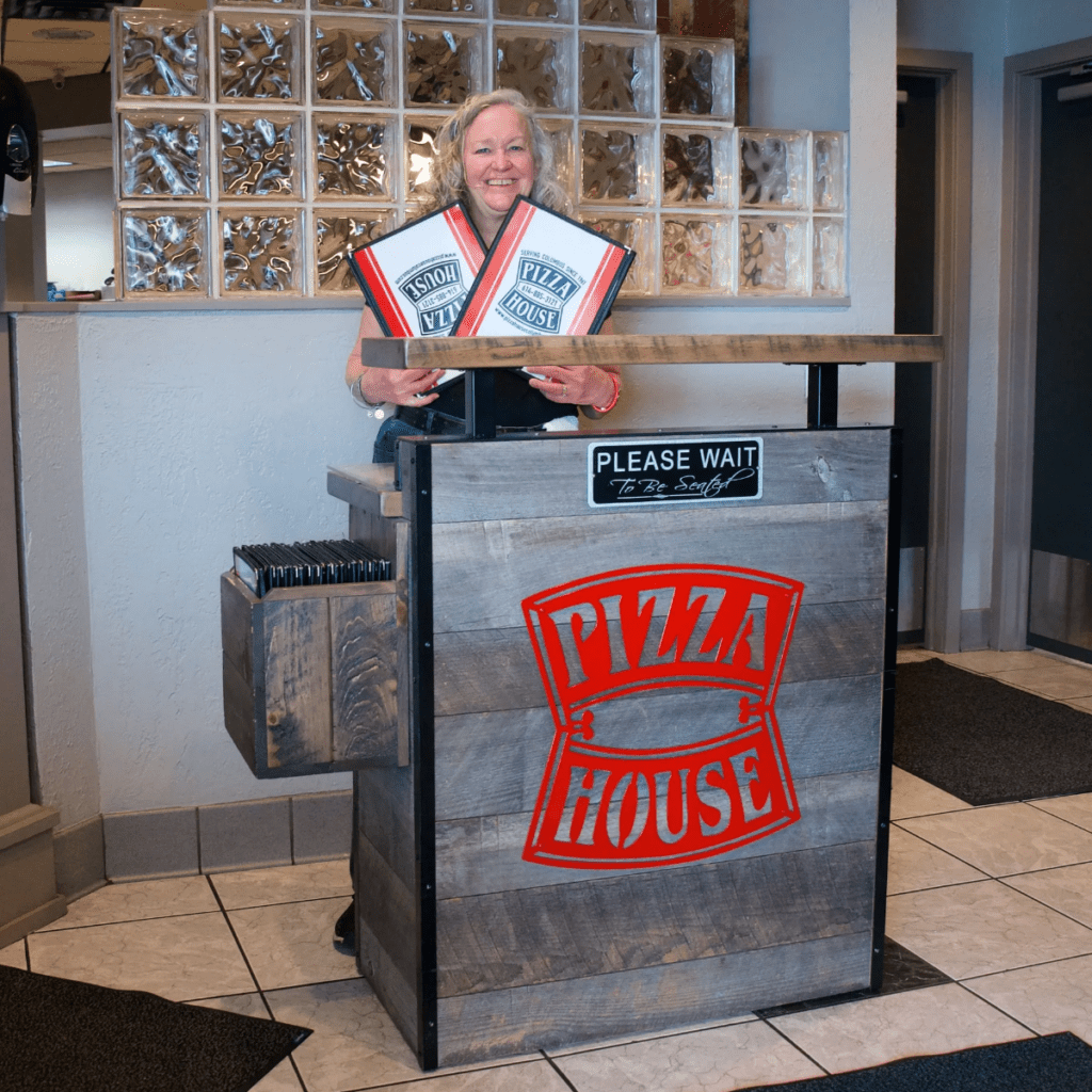 A smiling hostess stands at the entrance of Pizza House, holding menus. A "PLEASE WAIT" sign and the restaurant's logo are visible.