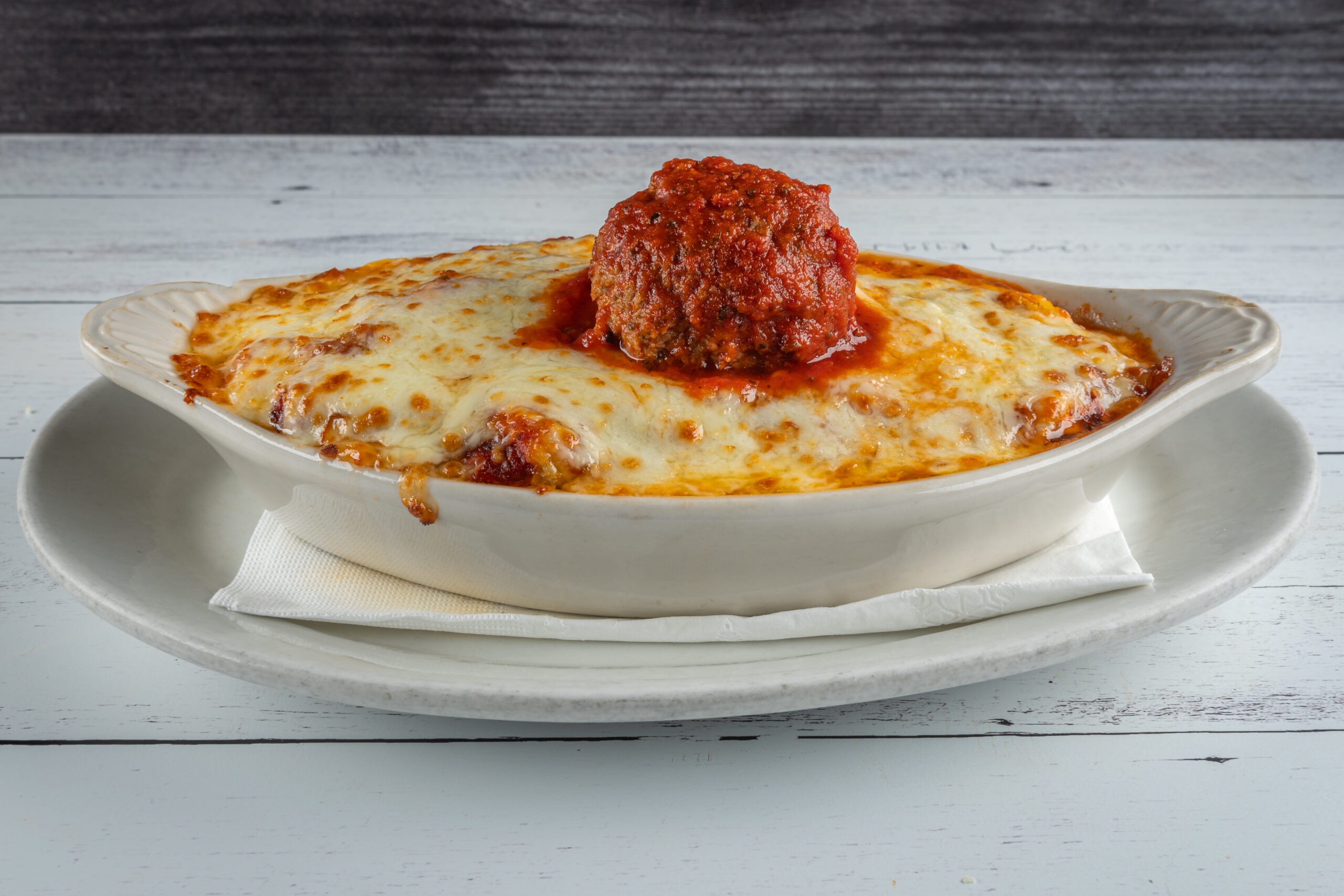 Baked pasta dish topped with melted cheese and a prominent meatball in marinara sauce, served on a white plate. Perfect for Italian cuisine enthusiasts.